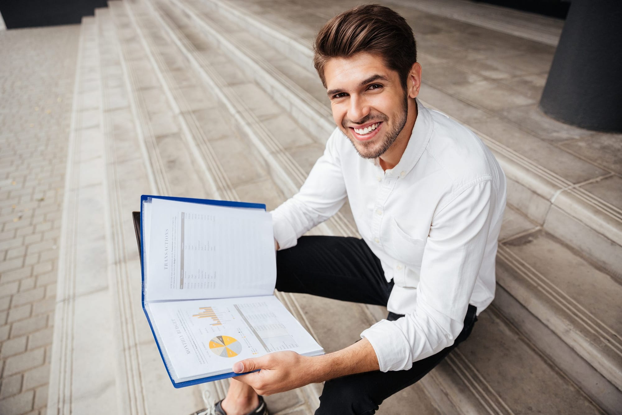 Cheerful young businessman sitting and reading business plan in folder
